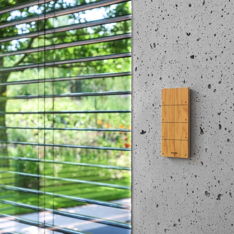 Panneau de commande Grenton en bois sur un mur en béton, à côté de stores extérieurs